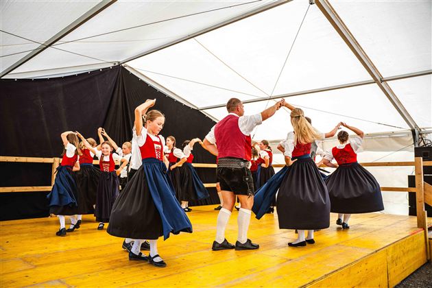 Eine Gruppe von Tänzern in traditionellen Trachten führt einen Volkstanz auf. Sie bewegen sich fröhlich auf einer Bühne in einem Zelt.