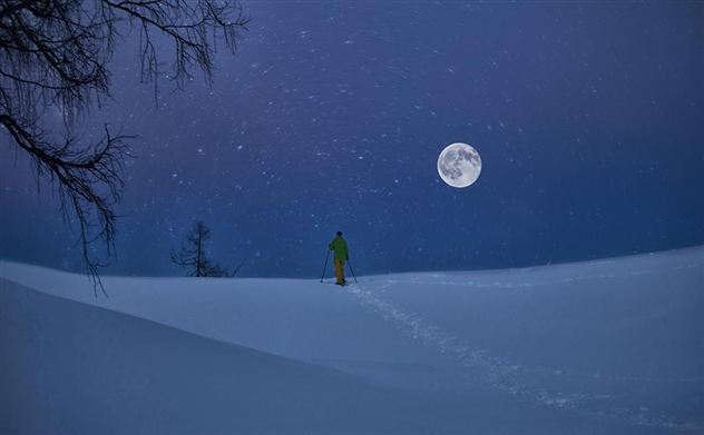 Ein skier steht in der verschneiten Landschaft unter dem Vollmond. Der Himmel ist dunkelblau und es schneit leicht.