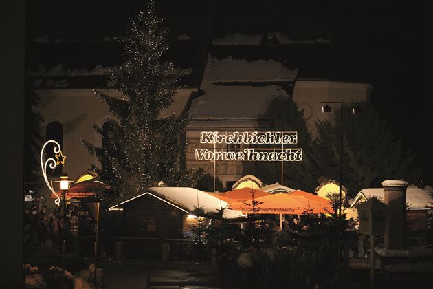 En festligt oplyst julemarked om natten. I baggrunden står et oplyst træ og en neonskilt med teksten "Kirchhöfler Vorweihnacht".