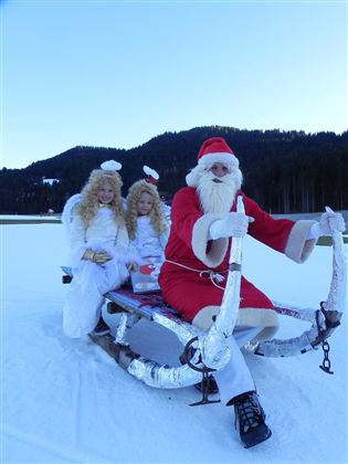 Ein Weihnachtsschlitten mit dem Weihnachtsmann und zwei Engeln auf einem verschneiten Feld. Im Hintergrund sind bewaldete Hügel und klarer blauer Himmel zu sehen.