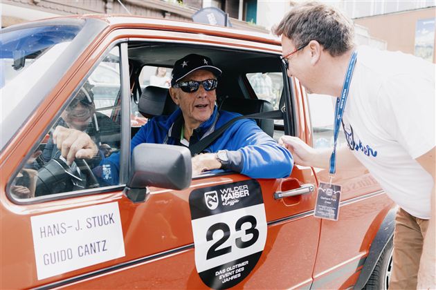 Ein älterer Mann sitzt in einem roten Auto und trägt Sonnenbrillen und eine Kappe. Ein weiterer Mann steht außen neben dem Auto und spricht mit ihm.