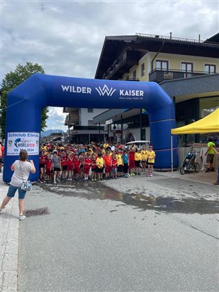 Eine Gruppe von Kindern steht unter einem großen blauen Startbogen, der den Schriftzug "Wilder Kaiser" trägt. Im Hintergrund sind Gebäude und eine gelbe Zeltstation sichtbar.