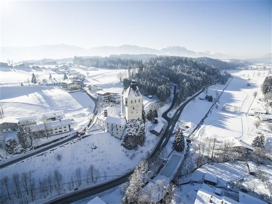 Een schilderachtig winterlandschap met een kasteel te midden van sneeuwbedekte velden. De omgeving is rustig en idillisch, omringd door bergen op de achtergrond.