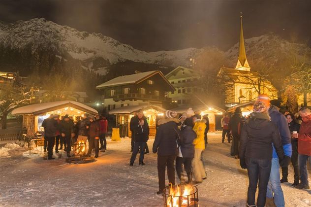 Ein festlicher Weihnachtsmarkt in den Bergen bei Nacht, umgeben von Schnee und beleuchtet von warmen Lichtern. Menschen versammeln sich um Feuerstellen, während im Hintergrund eine Kirche sichtbar ist.