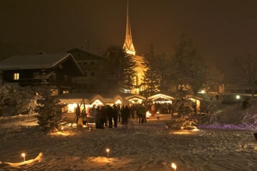 Eine zauberhafte Winternacht mit einem Weihnachtsmarkt. Menschen versammeln sich unter schneebedeckten Bäumen und um festlich beleuchtete Stände.