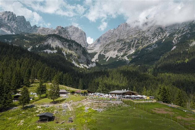 Eine malerische Berglandschaft mit hohen Gipfeln und dichtem Wald. Im Vordergrund ist eine gemütliche Hütte und eine Terrasse mit Gästen zu sehen.