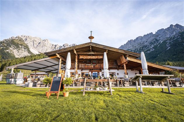 Ein traditionelles Alpenhaus mit großzügiger Terrasse und Sonnenschirmen. Im Hintergrund erheben sich majestätische Berge unter einem klaren Himmel.