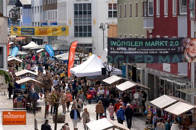 Eine lebhafte Fußgängerzone mit vielen Menschen und Verkaufsständen. Über den Ständen hängen bunte Banner, die auf den Wörgler Markt hinweisen.