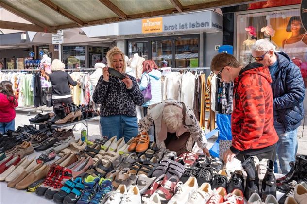 Ein gemütlicher Markt mit einer großen Auswahl an Schuhen und fröhlichen Menschen. Im Hintergrund sind Kleidung und andere Verkaufsstände zu sehen.