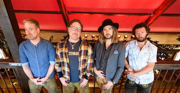 Eine Gruppe von vier Männern steht auf einer Tribüne. Sie tragen verschiedene Outfits und posieren vor einem roten Hintergrund.