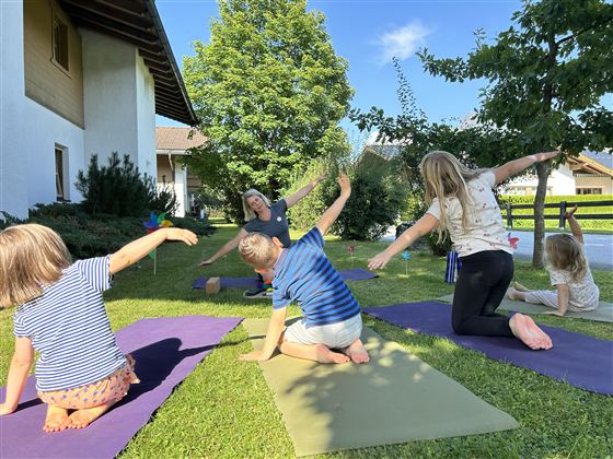 Een groep kinderen doet yoga buiten op groene matten. Op de achtergrond zijn een huis en bomen te zien.