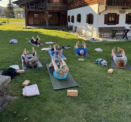 Eine Gruppe von Menschen macht Yoga draußen auf einer Wiese. Im Hintergrund sieht man ein traditionelles Holzhaus und einen klaren Himmel.