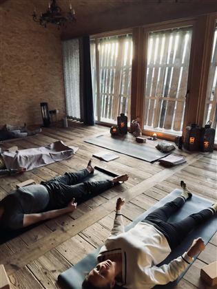 Ein ruhiger Raum mit Holzfußboden, in dem Menschen entspannt auf Yogamatten liegen. Sanftes Licht strömt durch die Fenster und schafft eine entspannende Atmosphäre.