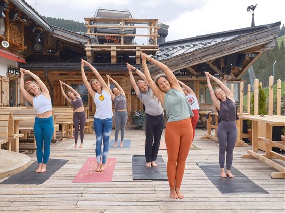 Gruppe von Menschen, die Yoga auf einer Holzterrasse macht. Im Hintergrund ist ein traditionelles Holzhaus zu sehen.