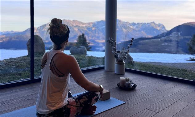 A person is sitting in a lotus position and meditating in a modern room with large windows. In the background, majestic mountains and a serene landscape can be seen.