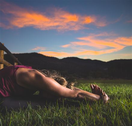 Een persoon doet yoga in het gras met een prachtige berglandschap op de achtergrond. De lucht is bij zonsondergang in zachte kleuren gedompeld.