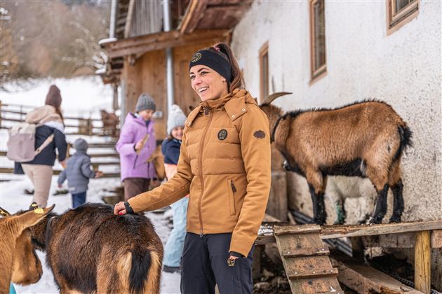 Eine Person mit brauner Jacke steht vor einem Stall und streichelt eine Ziege. Im Hintergrund sind weitere Personen zu sehen, die sich ebenfalls mit Ziegen beschäftigen.