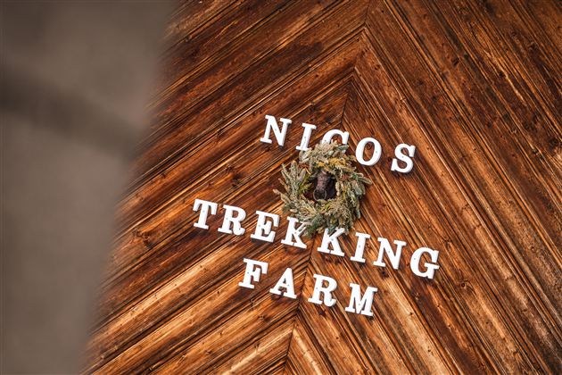 Ein Holzboden mit dem Schriftzug "Nicos Trekking Farm" und einem grünen Kranz. Die Atmosphäre ist rustikal und einladend.