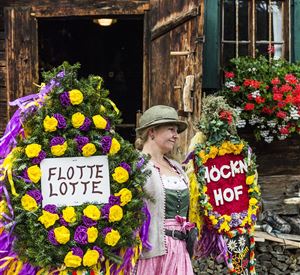 Eine Frau in traditioneller Tracht steht vor einem Holzhaus und trägt zwei festlich geschmückte Kränze. Die Kränze sind mit bunten Blumen dekoriert und tragen die Schriftzüge "FLOTTEN LOTTE" und "HÖCKEN HOF".