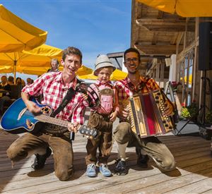 Zwei Musiker in traditioneller Kleidung spielen mit einem Kind. Sie stehen auf einer Terrasse mit gelben Sonnenschirmen im Hintergrund.