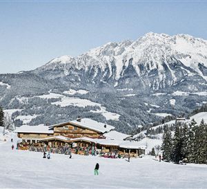 Eine malerische Winterlandschaft mit schneebedeckten Bergen. Im Vordergrund steht eine Berghütte, umgeben von Skifahrern und Tannenbäumen.