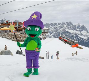 Een groene tovenaar in een kleurrijk kostuum staat in de sneeuw voor een berglandschap. Op de achtergrond zijn een skilerp en een berghut te zien.