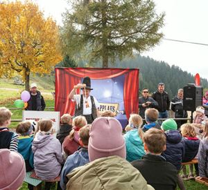 Eine fröhliche Kinderveranstaltung im Freien mit einem Zauberer auf der Bühne. Viele Kinder und Erwachsene schauen gespannt zu, während bunte Bäume im Hintergrund sichtbar sind.