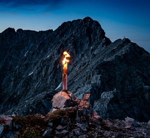Eine Fackel brennt auf einem Felsen in den Bergen. Im Hintergrund sind beeindruckende Berggipfel und ein dunkler Himmel zu sehen.