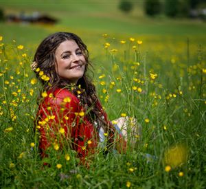 Eine lächelnde Frau sitzt in einem blühenden Feld mit gelben Blumen. Sie trägt ein rotes Kleid und genießt die Natur.