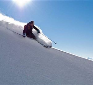 Ein Skifahrer fährt mit Schwung die verschneite Piste hinunter. Der Himmel ist klar und sonnig, und frischer Schnee wird aufgewirbelt.