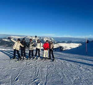 Five skiers are standing on a snowy slope. In the background, there are mountains and a clear blue sky.