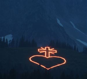 Een verlicht hart met een kruis in de bergen bij schemering. De lichten stralen in de donkere omgeving.