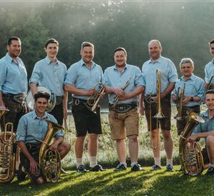 Eine Gruppe von Musikern steht im Freien und trägt traditionelle Trachten. Sie halten verschiedene Blasinstrumente und lächeln in die Kamera.