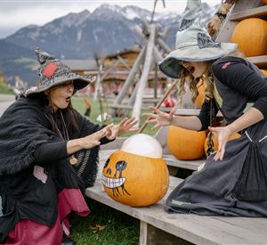 Two women in witch costumes are standing in front of a wooden shelf with pumpkins. They seem to be looking at a pumpkin with a face and appear to be excited.