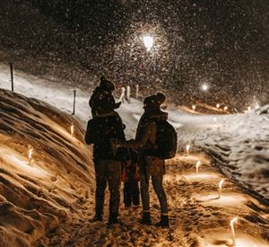 Een gezin wandelt 's nachts door de sneeuw terwijl de sneeuwvlokken vallen. De weg is omzoomd met lichten die een feestelijke sfeer creëren.