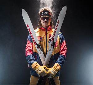 Een skier in een kleurrijke skijas houdt twee ski's in zijn handen. Hij draagt een zonnebril en heeft een sportieve haarstijl.