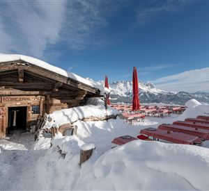 Eine rustikale Berghütte in einer verschneiten Berglandschaft. Rote Sonnenschirme stehen an den Tischen im Freien.