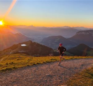 Eine Person joggt auf einem Pfad in den Bergen bei Sonnenuntergang. Im Hintergrund sind malerische Landschaften und ein farbenfroher Himmel zu sehen.