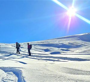 Three people are hiking on a snowy terrain. The sky is clear and the sun is shining brightly.