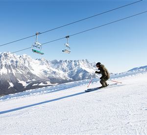 Een skiër skiet op een sneeuwbedekte piste met indrukwekkende bergen op de achtergrond. In de lucht zijn skiliften te zien.