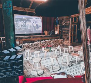 Ein gemütlicher Veranstaltungsraum mit Stühlen und einem großen Bildschirm. Der Raum hat eine rustikale Atmosphäre mit Holzelementen und einer Filmklappe im Vordergrund.
