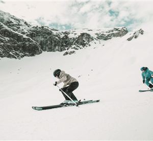Zwei Skifahrer fahren über einen schneebedeckten Hang. Im Hintergrund sind beeindruckende Berge und ein bewölkter Himmel zu sehen.
