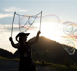 Eine Person steht im Sonnenuntergang und bläst Seifenblasen. Die Berge und der Himmel sind im Hintergrund gut sichtbar.