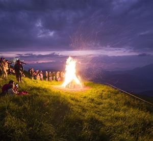 En gruppe mennesker står på en mark og observerer et stort bål under en overskyet himmel. Funken stiger op i natten, mens scenen giver en festlig stemning.