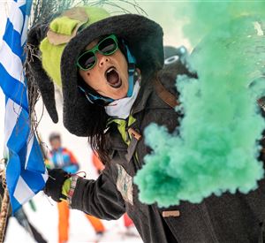 A person is wearing a large hat and sunglasses and is firing a green smoke flare. In the background, people can be seen skiing.
