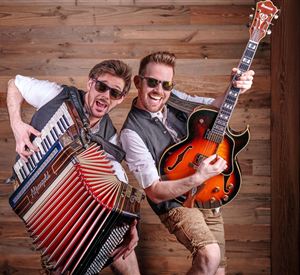 Zwei Musiker posieren fröhlich vor einer Holzwand. Einer spielt das Akkordeon und der andere hält eine Gitarre.