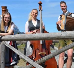 Three musicians are standing on a wooden bridge in the mountains. They are playing double bass and accordion and are wearing traditional costumes.