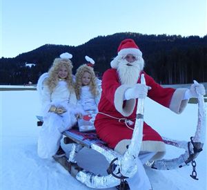 A Christmas sleigh with Santa Claus and two angels on a snowy field. In the background, there are forested hills and a clear blue sky.