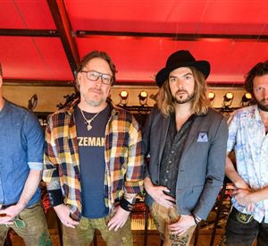 Eine Gruppe von vier Männern steht auf einer Tribüne. Sie tragen verschiedene Outfits und posieren vor einem roten Hintergrund.