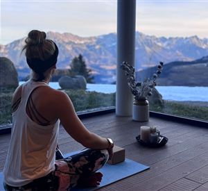 Eine Person sitzt im Lotussitz und meditiert in einem modernen Raum mit großen Fenstern. Im Hintergrund sind majestätische Berge und eine ruhige Landschaft zu sehen.
